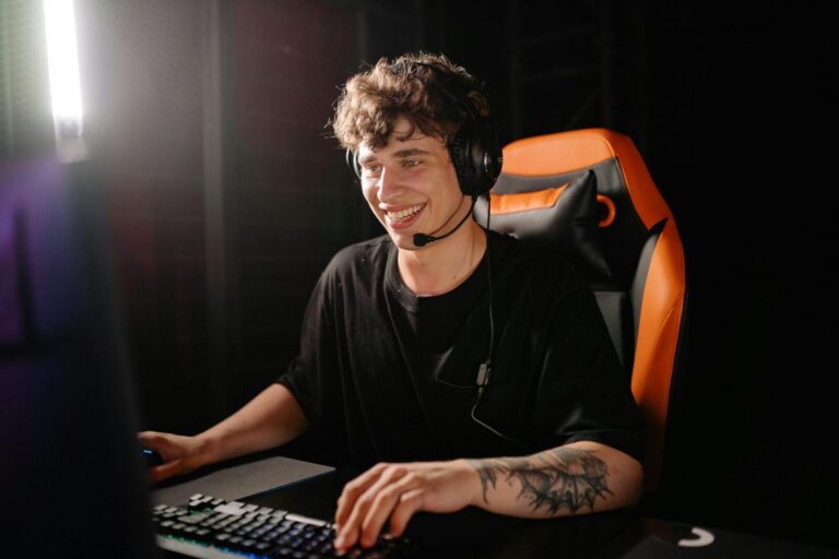 A smiling young man enjoys PC gaming on a computer with headset in an indoor dark setting.